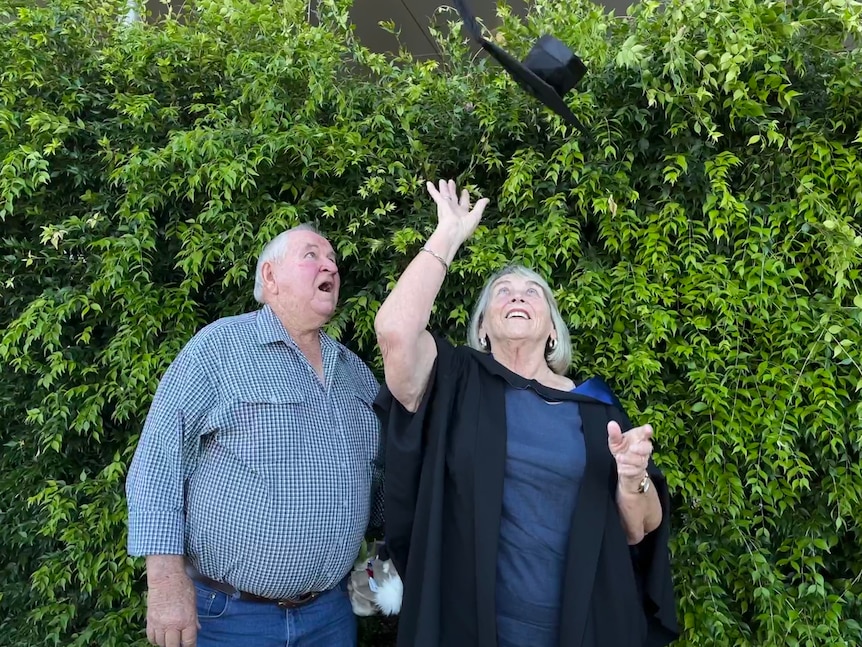 An older couple standing in front of a green hedge, both smiling as the woman throws a graduate's hat into the air.