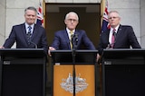 Three men in suits stand next to each other at three podiums, the central podium is decorated with the Australian crest