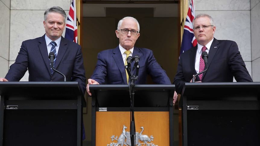 Three men in suits stand next to each other at three podiums, the central podium is decorated with the Australian crest