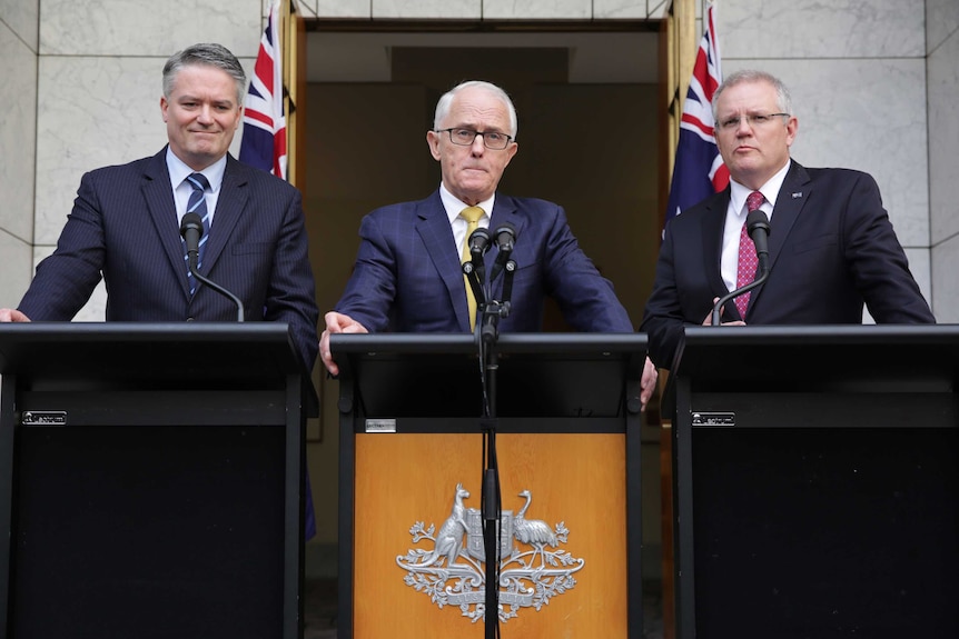 Three men in suits stand next to each other at three podiums, the central podium is decorated with the Australian crest