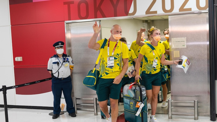 Australian athletes in green and gold tracksuits and face masks wave at an airport terminal 