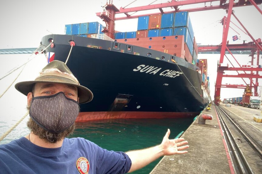 A man in a face mask holds out his arm in front of a large cargo ship docked in a port