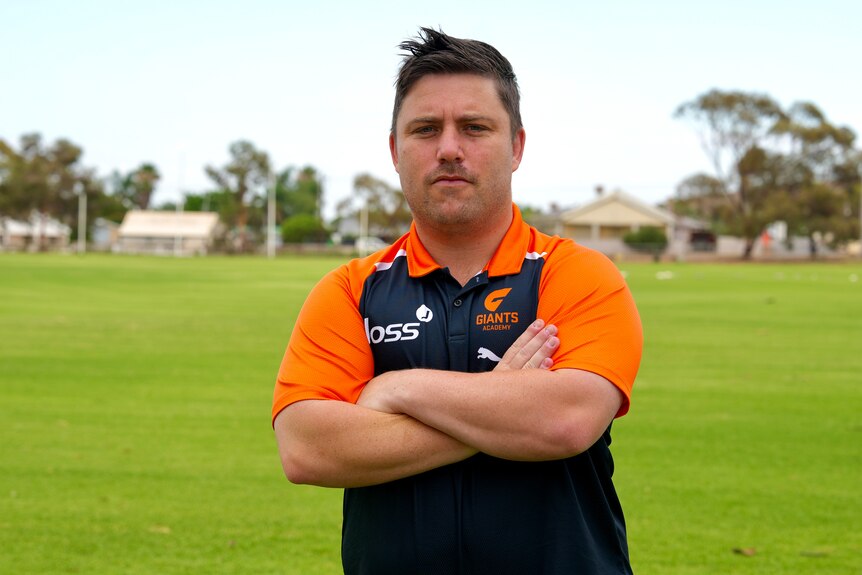 A white man with short brown hair wearing a charcoal and orange polo shirt with his arms crossed