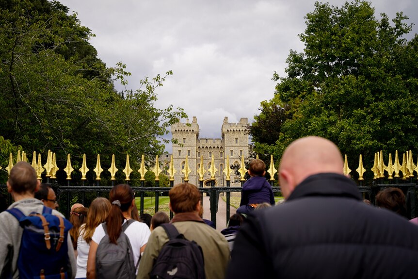 A crowd of people gather outside gold, wrought-iron gates. In the distance, Windsor Castle is visible