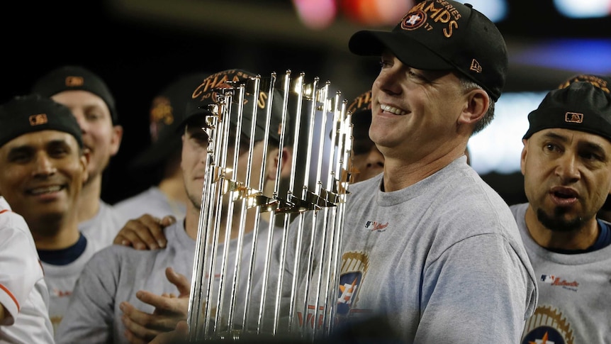 AJ Hinch smiles while holding the World Series trophy, surrounded by players from the team.