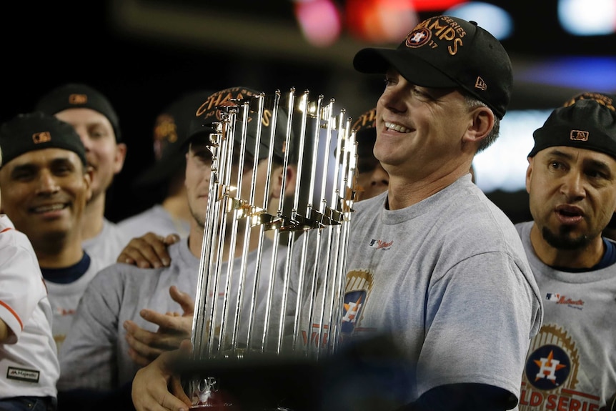 AJ Hinch smiles while holding the World Series trophy, surrounded by players from the team.