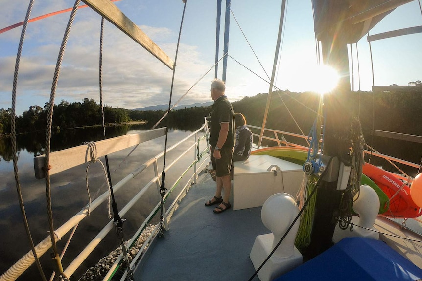 Trevor Norton standing on the deck at sunset, susnhine lights the mast and rigging.