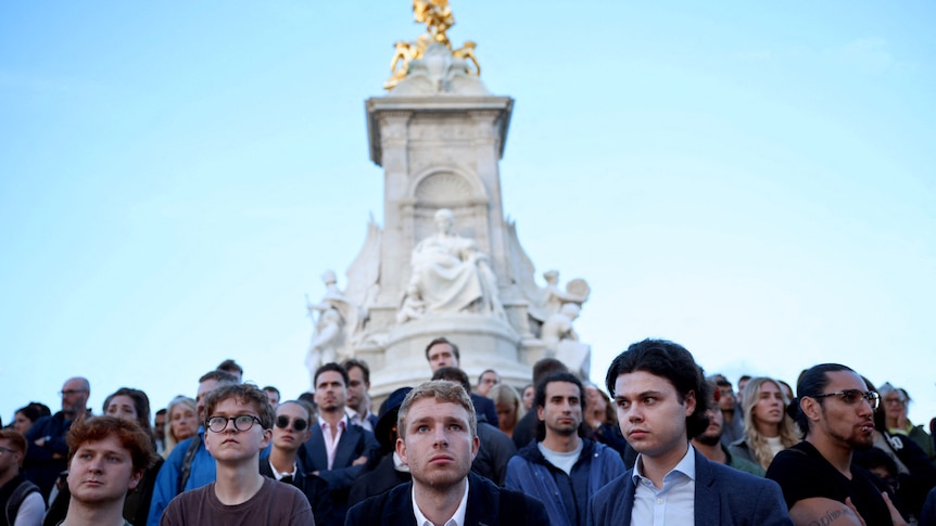 A group of people standing in front of the Victoria Memorial outside Buckingham Palace looking sombre