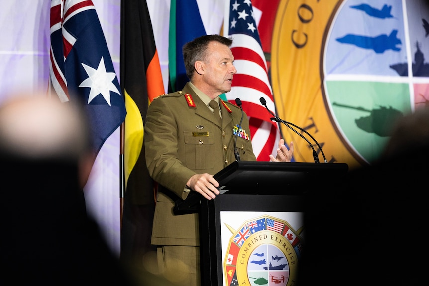 A man in Australian military uniform speaks sternly at a podium in front of Australian and US flags