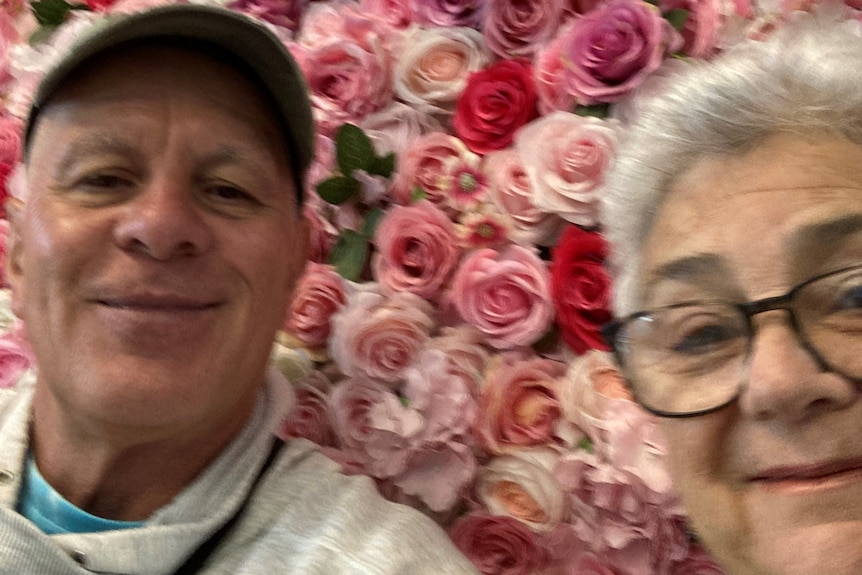 Older man and woman take selfie in front of wall of pink roses.