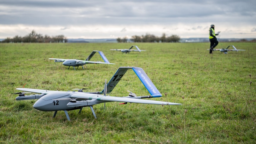 Three drones are positioned in the middle of a green field, against a grey sky. A person in the background holds a remote