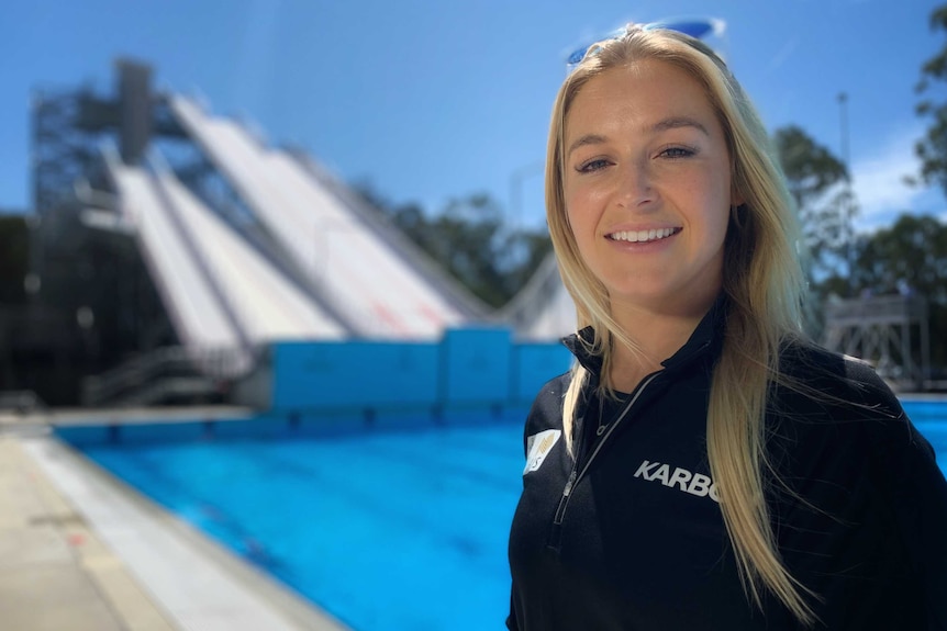 A young blonde woman in black sports tracksuit smiles at camera with pool and slide behind her.
