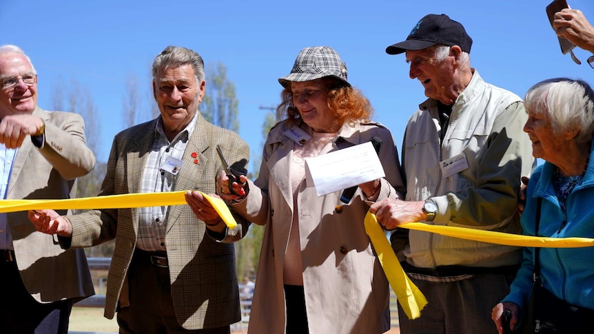An older woman standing in the middle of a row of five older people cuts a ribbon that they are all holding.