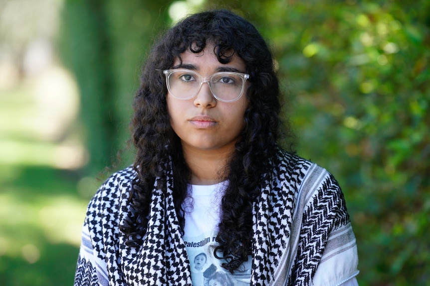 A young woman with curly black hair and glasses looks serious.