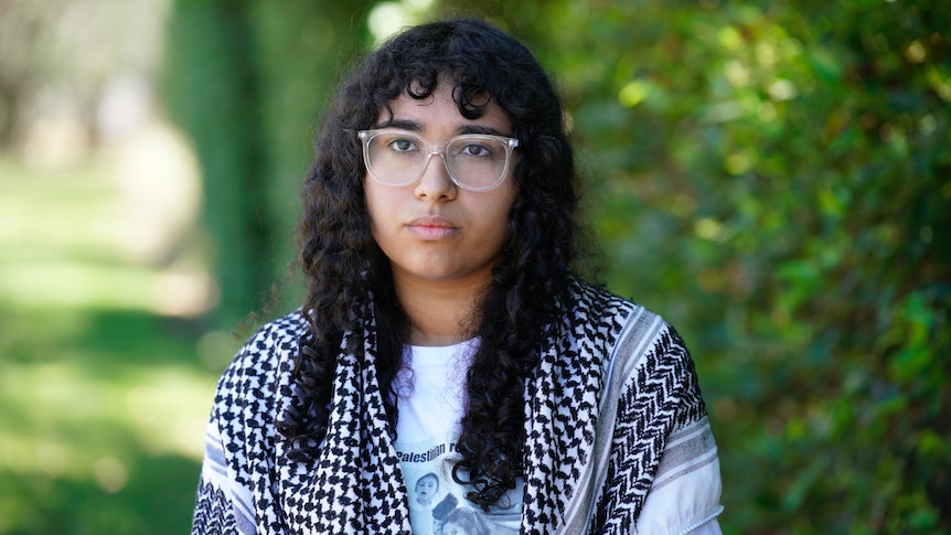 A young woman with curly black hair and glasses looks serious.