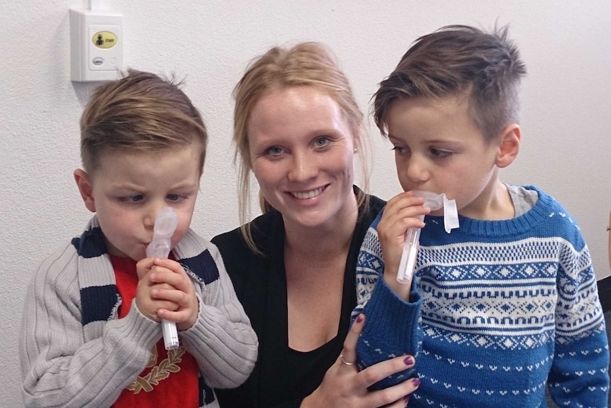 A woman crouches between two boys with plastic tubes in their mouths.