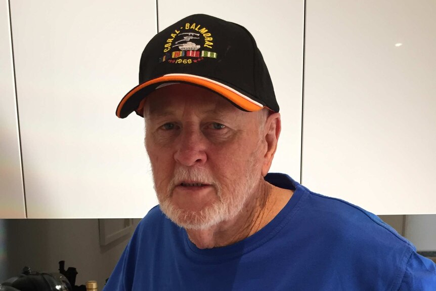 A man wearing a cap with Coral Balmoral on it standing in a kitchen.