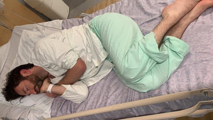 Taken from above, a man lying on his side on a hospital bed. He is sleeping with his hands curled under his chin.