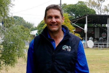 man in blue shirt and navy vest standing in a yard with a tree and shed in the background