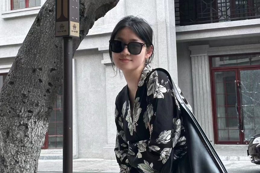 A woman with black sunglasses standing on a street smiling and carrying a black bag
