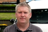 A man in a grey t-shirt stands outside with a white van behind him.