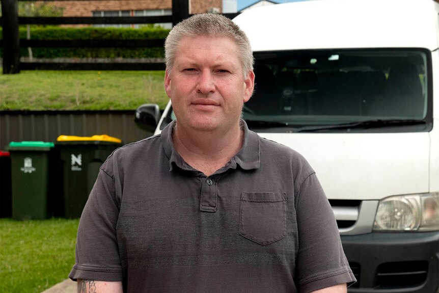 A man in a grey t-shirt stands outside with a white van behind him.