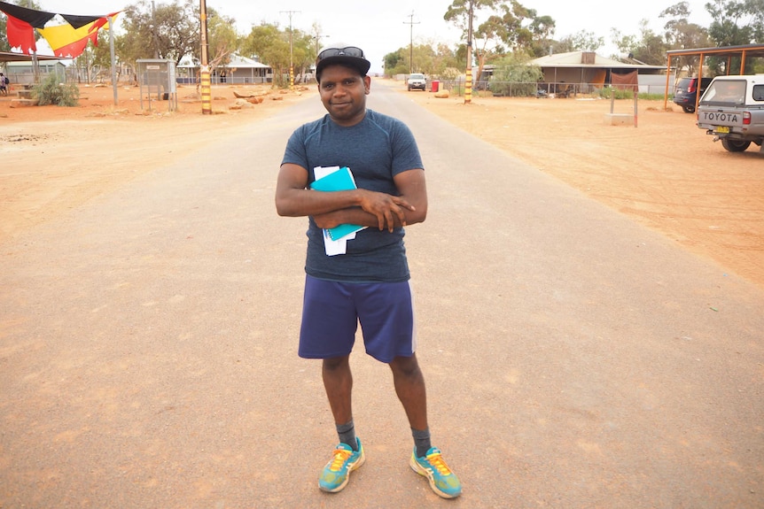 A man stands in the middle of a street with a cap on holding paper near his chest and smiling