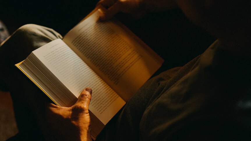man's hands holding open hard back book on his lap, in late afternoon light