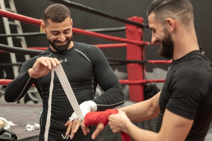Two men smile as they tape up their hands to go into an MMA training fight.