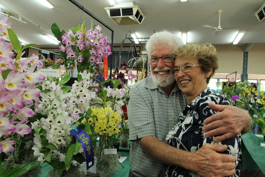 A man hugs a woman standing beside flowering orchid plants