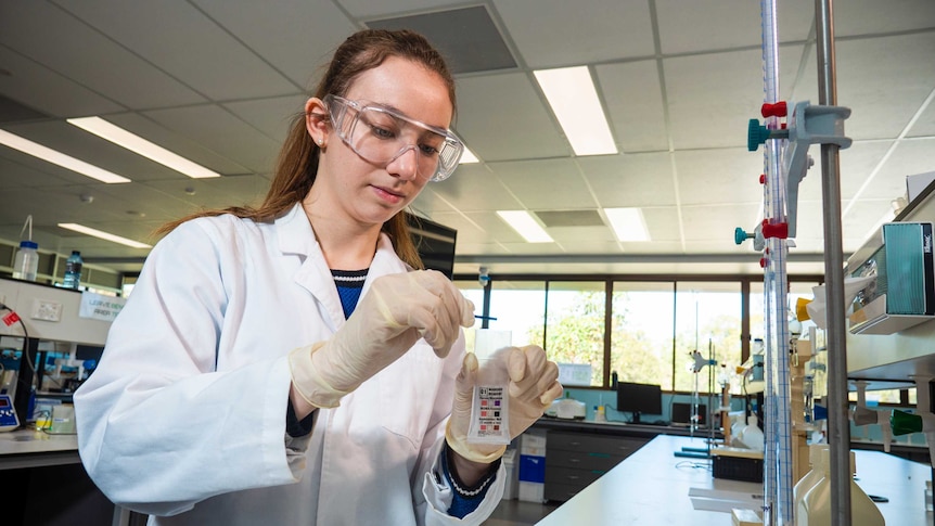 Jasmine White in a lab coat and goggles and gloves and holding a test tube