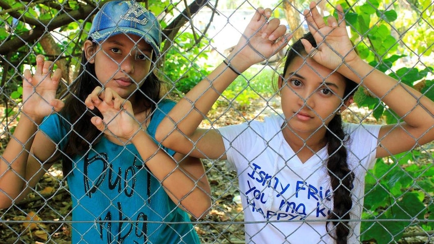 Two young refugee girls stand behind a barbed wire fence in nauru.
