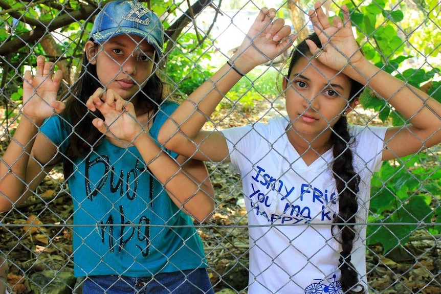 Two young refugee girls stand behind a barbed wire fence in nauru.