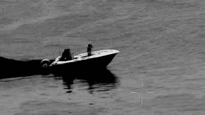 A black and white aerial image of a recreational vessel with civilian passengers on board before being rescued.