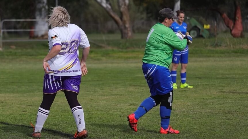 Goalkeeper with a green jersey claims a ball in the field as a player in purple shorts, lavender tee has back on camera.