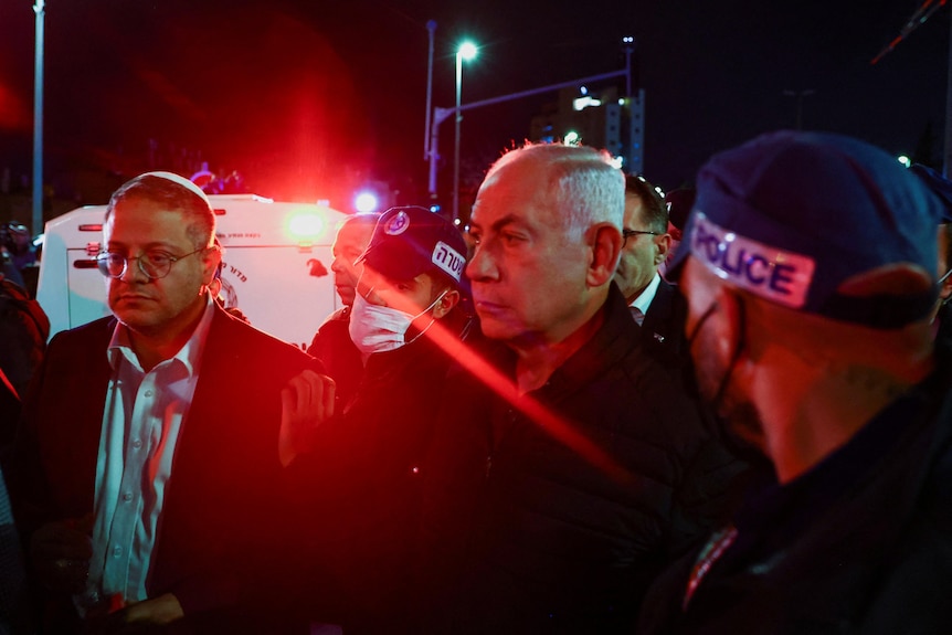 Two senior politicians are surrounded by police and illumated by emergecy services lights as they visit a crime scene at night.
