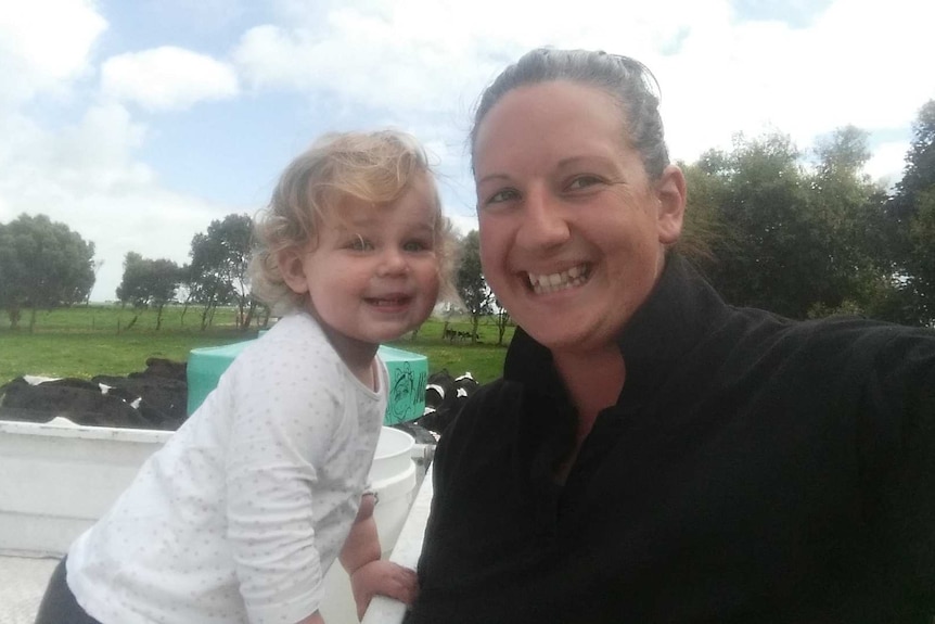 A blonde toddler stands on a ute tray in a paddock next to her mum, with black and white dairy cows in the background.