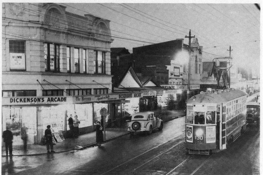 Black and white photo of a street with a tram going past and an old car