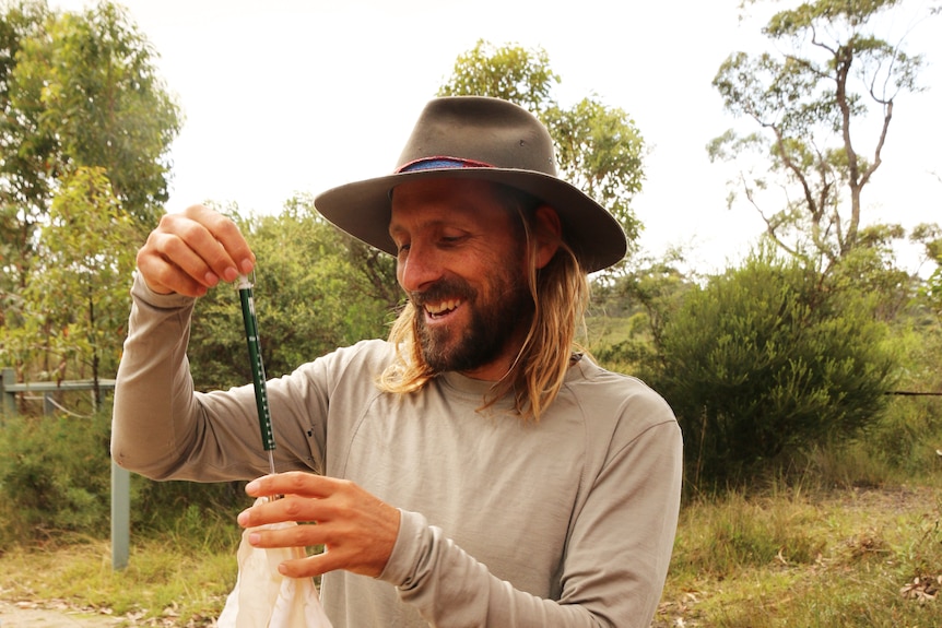 David Bain wears a wide brimmed hat and smiles as a holds a green plastic object.