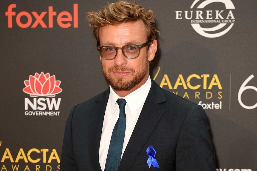 A full length shot of Simon Baker with a blue ribbon pinned on his pocket.
