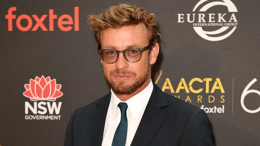 A full length shot of Simon Baker with a blue ribbon pinned on his pocket.
