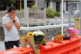 A mourner stands behind orange barriers with flowers placed on top. He has his face in his hands and is wearing a grey shirt.