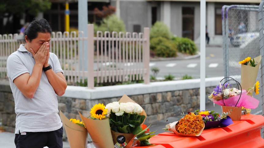 A mourner stands behind orange barriers with flowers placed on top. He has his face in his hands and is wearing a grey shirt.