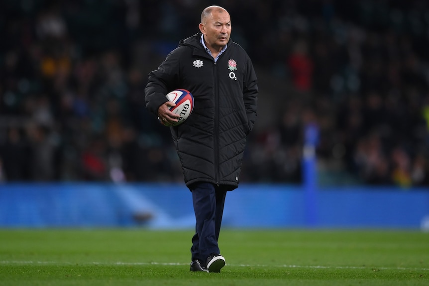 England men's national rugby union coach stands on a field holding a ball with his right arm.