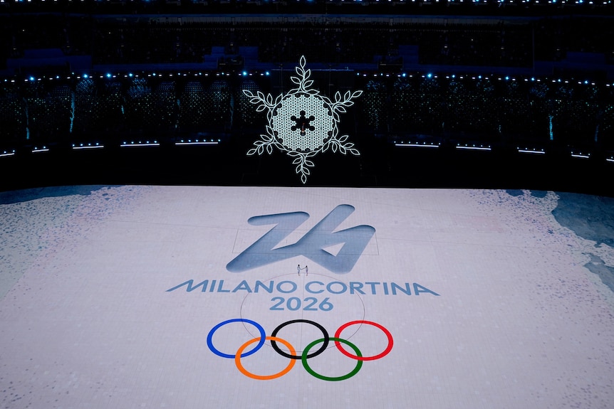 A giant snowflake hangs in the air above the stadium floor with an image of an Olympic logo "Milan-Cortina 2026".
