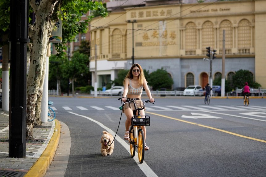 A young Chinese woman rides her bike down a city street, with a dog trotting along beside her