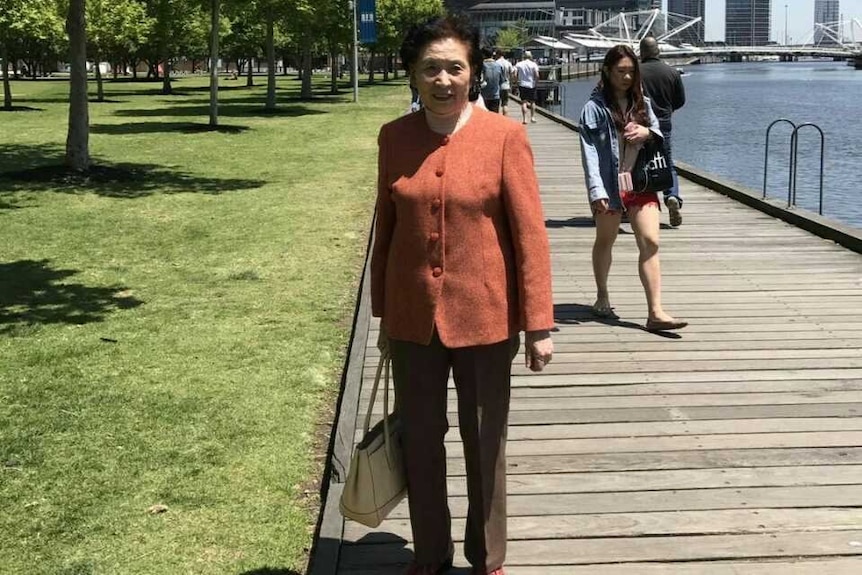 A woman in a red coat holds her handbag in her right hand as she stands on a boardwalk, with large glass buildings seen behind.