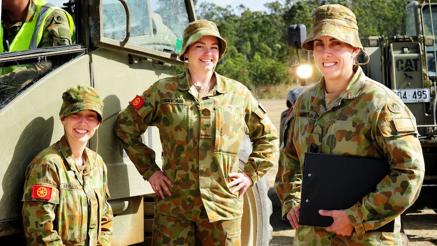 Three female soldiers stand near a truck smiling. They wear dark green and brown army fatigues.