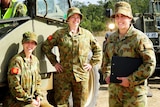 Three female soldiers stand near a truck smiling. They wear dark green and brown army fatigues.