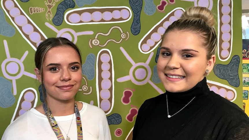 Sisters, Merinda and Sophie Dryden, standing in front of a wall painted in an Aboriginal style